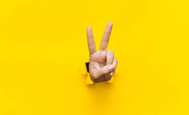 a vitória assina gesto de mão de dois dedos em fundo amarelo com orifício de papel rasgado e espaço de cópia. é um conceito ok. - two fingers - fotografias e filmes do acervo