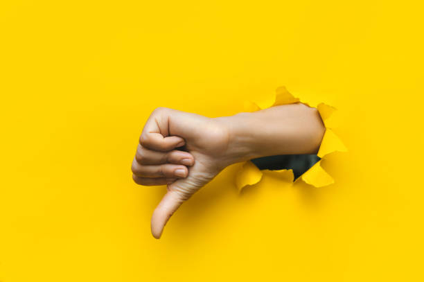 istock Hand showing a thumb down through ripped hole in bright yellow paper background. Concept of dislike and disapproval gesture. 1359581490