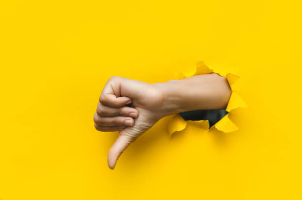 Hand showing a thumb down through ripped hole in bright yellow paper background. Concept of dislike and disapproval gesture. stock photo