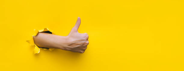 The right female hand shows a thumb up gesture (like). Torn hole in yellow paper. The concept of positive attitude, approval and praise. Copy space. stock photo
