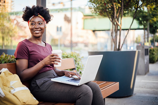 Portrait of a beautiful young African American woman sitting on the street, using her laptop and enjoying coffee.