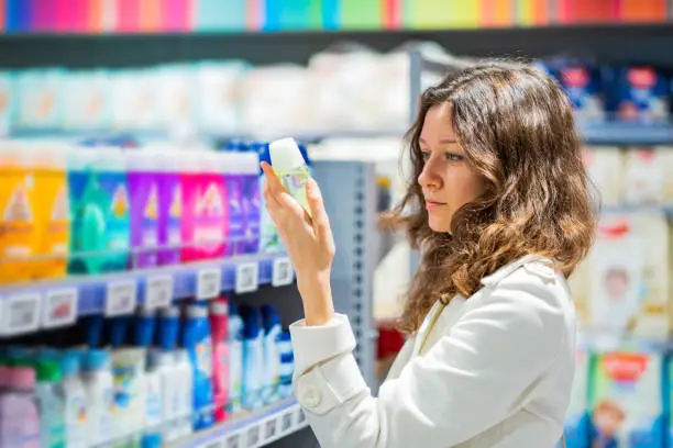 Brunette woman with long curly hair wearing white coat reads instruction on deodorant package standing in household goods store