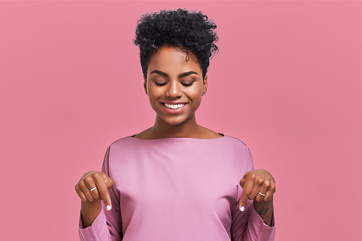 Funny surprised African American female looks with joyful eyes down and indicates as shows something, sees comic things, isolated over pink wall. Positive amazed young woman poses in studio.