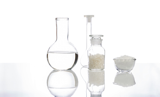 White flake chemical in glass container place next to Sea Salt in Chemical Reagent Bottle Glass, Crystal clear liquid in Flat Bottom Flask and Volumetric flask. Side View