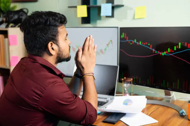 Worried trader praying god to make profit or stock market to go up in front of charts on computer screen - Concept of risk in crypto trading and investing on equity shares