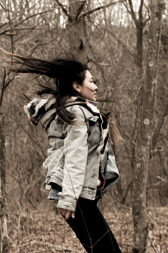 A photo from the side of a young Japanese woman with long hair in a denim jacket jumping happily in a natural forest.