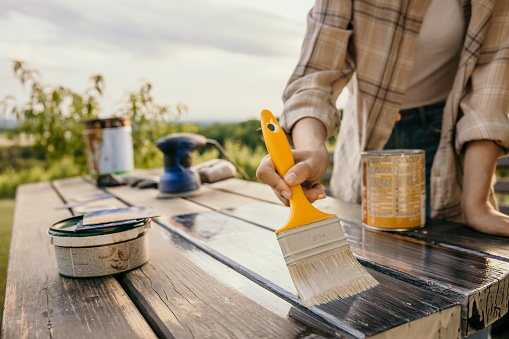 Adult woman painting a old wooden table with a yellow brush in the garden on a cloudy day,no face,varnish can and tools in background,home improvement,horizontal