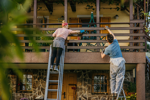 Close-up adult couple man and woman in their 30s on two ladders wearing old clothes for renovation,painting the wooden balcony of their house with brushes to apply brown color for protection against climate and weather,concentrated,home improvement,horizontal