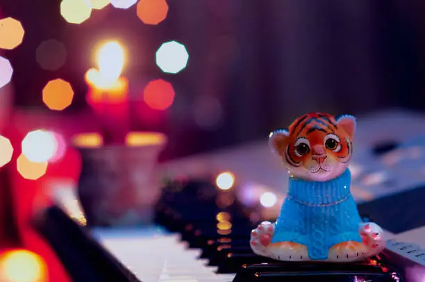 Photo of A figurine of a toy tiger cub in a blue sweater sits on the keys of a musical instrument. A candle is burning in a candlestick. The glow of the New Year's garland. Christmas tree decoration.