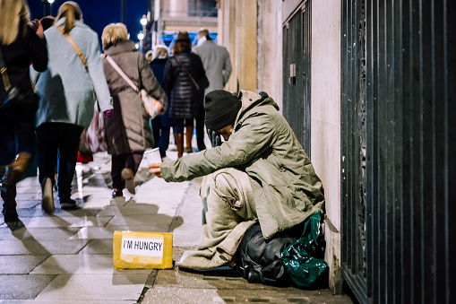 London, UK - 10 December, 2021: Color image depicting a homeless man begging for money on a busy city street. Crowds of people walk past him, ignoring his presence.