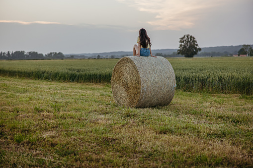 Young woman with long brown hair sitting on a hay ball in the stubble field in front of a grown field,enjoying the nature during evening time in the summer,view from the back,hills in background,horizontal