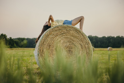 Close-up of a natural looking young woman with long brown hair,eyes closed touching her head while lying on a perfectly formed hay ball in the stubble field,view from the right side,entire body,summertime,evening,horizontal
