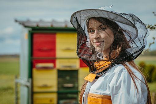 Close-up of a young beekeeper woman in her 20s wearing a white orange protective suit,looking and smiling at the camera,face and upper body parts,beehive in the background,horizontal