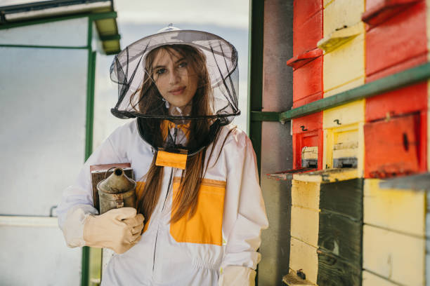 Young attractive beekeeper woman in her 20s with long dark blonde hair and white protective suit,holding a used look metal bee smoker under her arm smiling and looking at the camera,colorful beehive next to her,horizontal