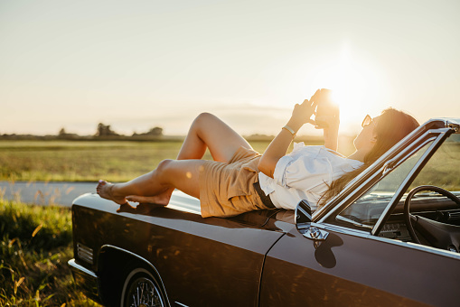Young woman with fancy clothes,long brown hair and sunglasses,looking at her smartphone while lying on the hood of her beautiful brown convertible car during sunset,sunbeam,entire body,fancy clothes,front of the car,horizontal