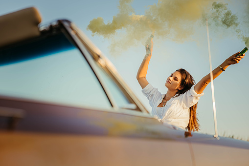 Close-up of a young attractive woman with brown hair,standing in front of her brown vintage convertible with her arms up in the air holding color bomb smoke spray in her hands,low angle,relaxed face,enjoying the moment,horizontal