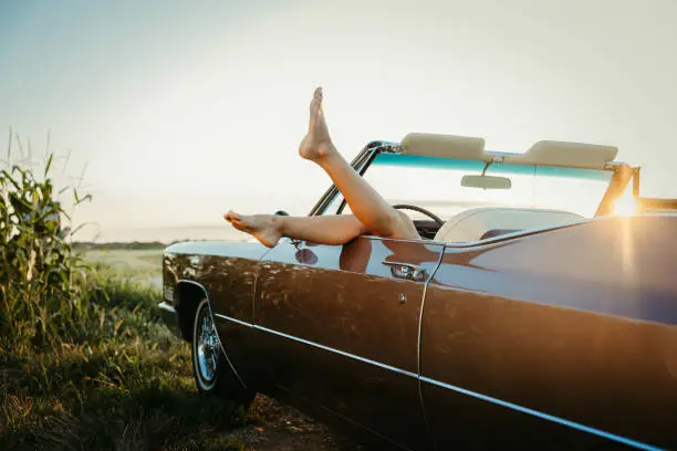 Photo of Beautiful brown convertible car parked next to a field with barefoot legs of a woman