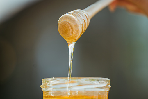 Close-up of a wooden honey dipper with yellow golden bee honey on it, flowing back down in a honey glass,horizontal