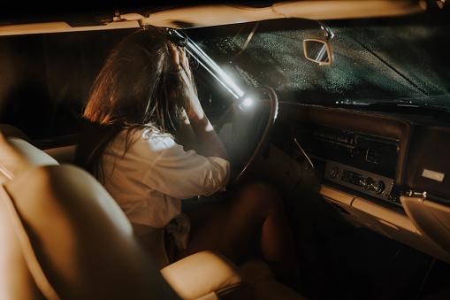 Young woman with long brown hair caused an accident while driving a vintage convertible during nighttime,rain on the windshield,woman holding her head while she leans on the steering wheel,view from back seat,horizontal