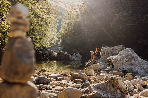 Adult woman with short brown hair sitting on a rock next to the lake reading a book,sunbeam ant plenty of rocks next to the lake,relaxed,in gorenjska,Slovenia,horizontal