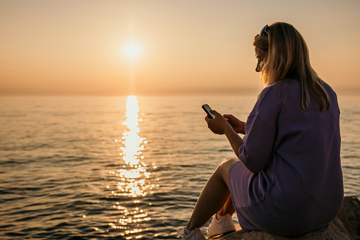 Adult woman in her 30s with purple dress and blonde long hair checking distracted her smartphone while sitting on a rock next to the sea horizon with sun and reflection in the ocean in background,view from the back,piran Slovenia,horizontal