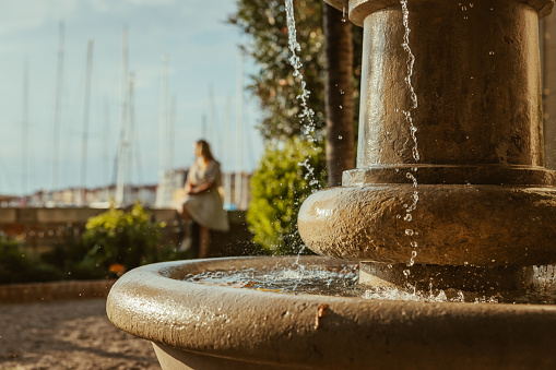 Old historic fountain with water drops,next to piran harbor. Out of focus adult woman in background sitting a wall looking at the harbor,daylight,close-up,Slovenia ,horizontal