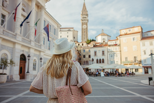 Blonde adult woman with long blonde hair wearing a white hat walking in the center of piran,Slovenia with cathedral St. Georg tower in background and tourist cafe in background,view of her upper body from the back,horizontal