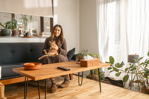 A pregnant woman is sitting on sofa and spending time and playing with her ginger cat in the living room at home.