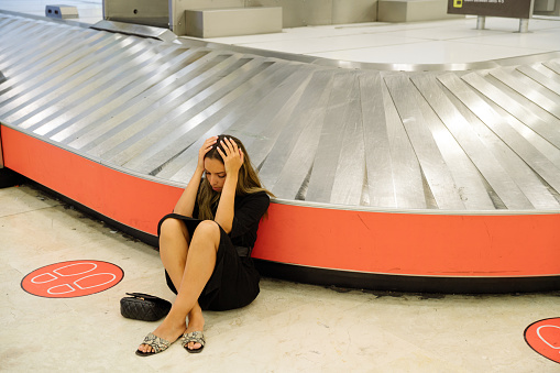 Worried female tourist sitting on floor near conveyor belt in airport and touching head because of lost suitcase