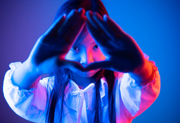 Asian child girl makes a triangular hand gesture and peeks through her hands in the dark with blue and red lights.