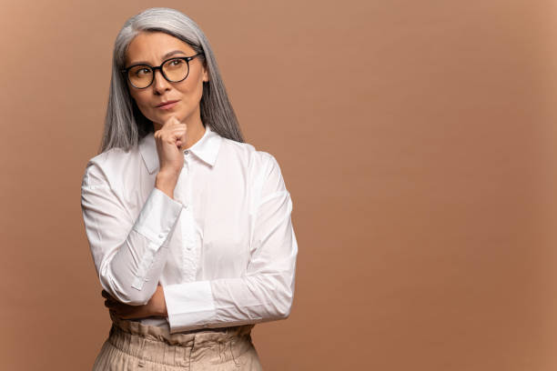 donna matura intelligente e riflessiva che tiene il mento e pondera l'idea, prende decisioni difficili, sembra incerta dubbiosa. studio interno girato isolato su beige - curiosità foto e immagini stock