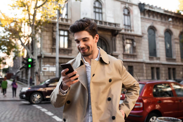 istock Young attractive smiling man in trench coat happily using cellphone walking through street 1359694234