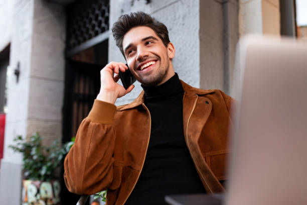 istock Young handsome casual man happily talking on cellphone while working on laptop in cafe on street 1359695925