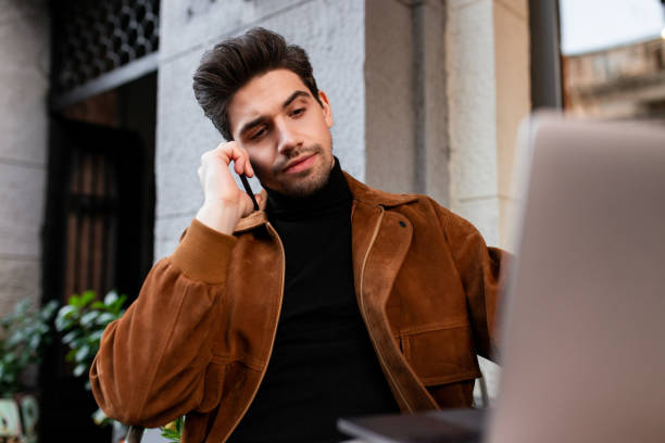 istock Young handsome man talking on cellphone confidently working on laptop in cafe on street 1359695932