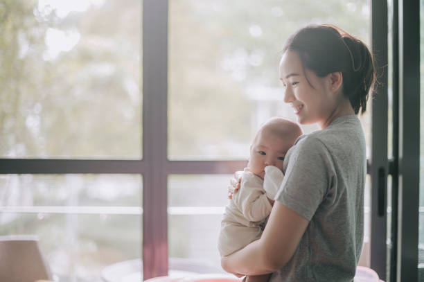 アジアの中国の母親は、バックライトの日光と朝のボンディング時間にベビーベッドから彼女の男の子を運びます - 空気感 ストックフォトと画像