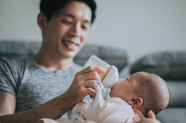 アジアの中国の若い父親は、週末にリビングルームでミルクボトルで彼の男の子の息子を養います - 動物の摂食 ストックフォトと画像