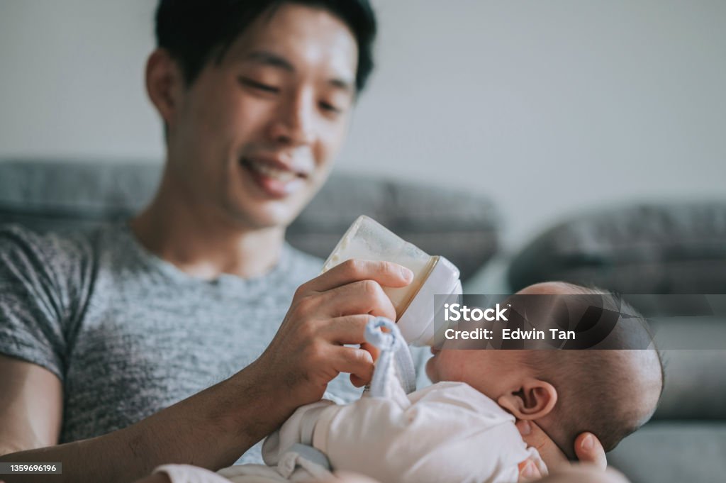 Asian Chinese young father feeding his baby boy son with milk bottle at living room during weekend Baby - Human Age Stock Photo