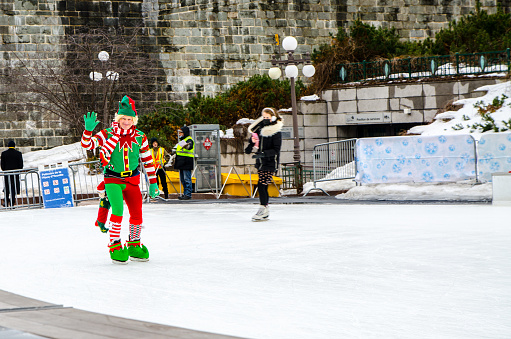 Man disguised in Christmas elf and ice skating on the rink in Old-Quebec during winter day on Place D'Youville.\nHe is waving at kids outside the rink