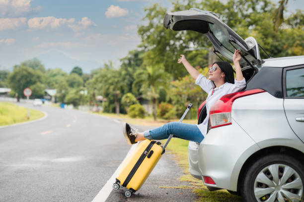 une touriste asiatique heureuse porte des lunettes de soleil assise sur une voiture à hayon avec des bagages jaunes pour s’amuser dans le parking. - location de voitures photos et images de collection