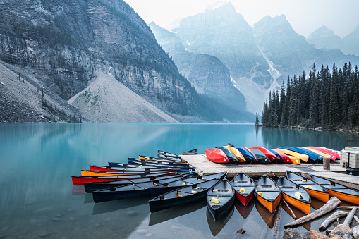 Colourful canoes provide a stark contrast to the still and foggy Moraine Lake at blue hour, in the distance, mountains tower overhead.