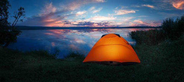 Internally lit orange tent on shore of lake under dramatic sunset sky