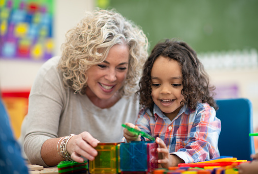 A preschool teacher leans in beside a little boy sitting at a table working on a building project, as she asks him about the structure.  The little boy is dressed casually and focused on his task.