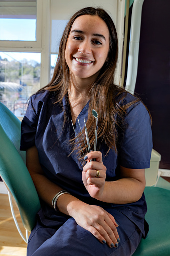 Front view of professional female dentist keeping dental tools, looking at camera and smiling in clinic. Pretty doctor posing while working in dentist office