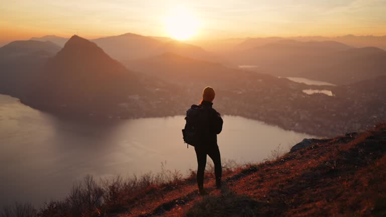 Female hiker stands at viewpoint with arms wide open, enjoying view of sunset, lake and city below