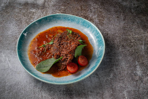 Beef stew with broth and herbs in a blue plate with spices and black pepper against a gray stone table