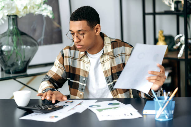 istock Focused modern hispanic young man with glasses, office worker or freelancer, sitting at his work desk, views graphs, analyzes reports and profit, works with documents in the office 1359746884