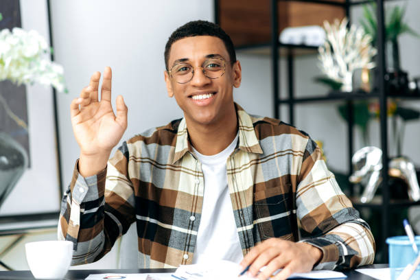 istock Distant communication. Charismatic friendly hispanic guy in glasses and in a casual shirt, sits at his work desk, looks at the camera, communicates on a video call, greets the interlocutor, smiles 1359746889