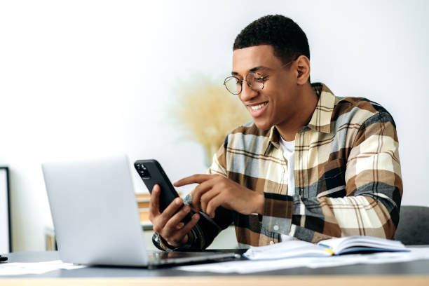 Satisfied latino modern guy with glasses, sitting at his desk, using his smartphone, texting with friends on social networks, browsing the Internet, writing email, makes an online order, smiling stock photo