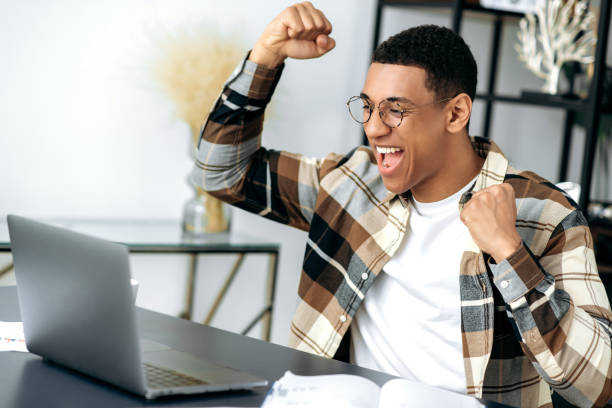 istock Happy joyful successful hispanic guy with glasses, stylishly dressed, employee, freelancer or student sits at work desk, gesturing with his hands rejoicing in victory, success, good deal, big profit 1359746915