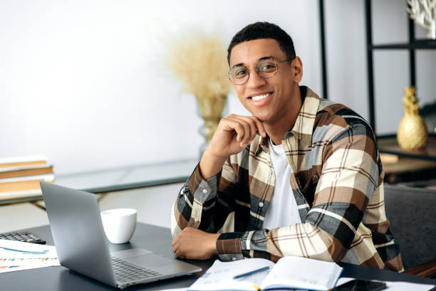 istock Portrait handsome confident successful young mixed race latino man, freelancer or manager, wearing shirt and glasses, sits at work desk with laptop, looking at the camera and smiling friendly 1359746918